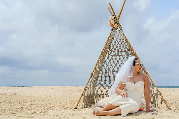 The bride in a white dress and veil sits on the sand in front of a macramé teepee.
