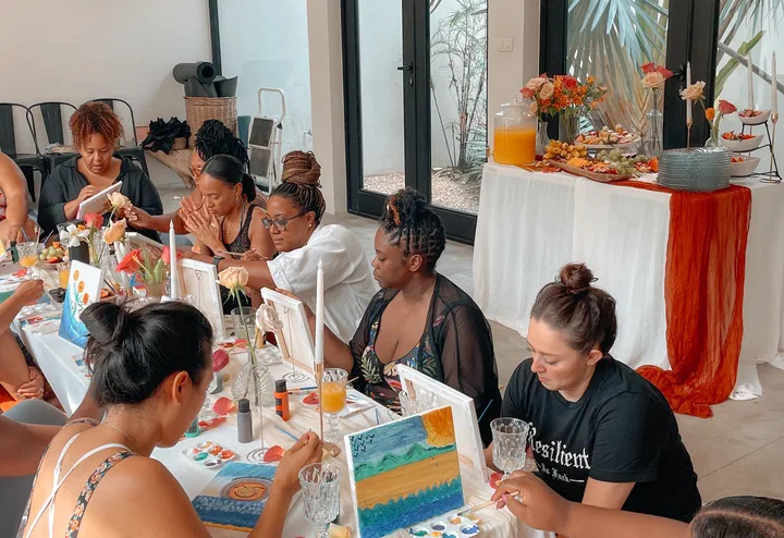 Women paint canvases at a long table in a bright studio, with a buffet of fruit and juice behind them.