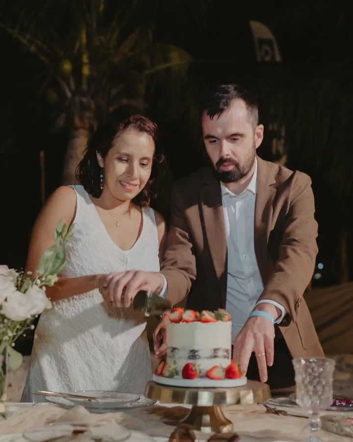 Una pareja corta juntos un pastel de fresas en una mesa de celebración con velas y flores blancas.