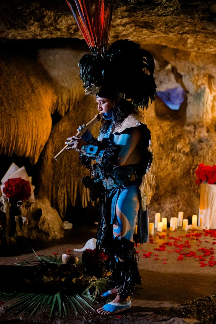 Un músico con atuendo de estilo maya y penacho de plumas toca la flauta en un cenote de caverna iluminado con velas.