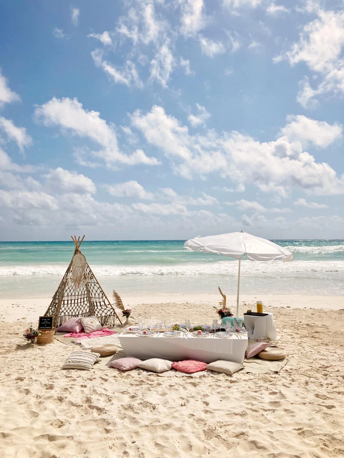 Low white picnic table with pink and white cushions, a teepee and a beach umbrella on the sand