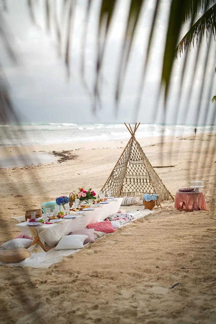 Beach teepee picnic with cushions and a low table, framed by a palm frond
