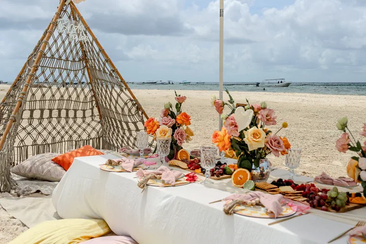 Long beach picnic table with orange and peach flowers beside a macramé teepee