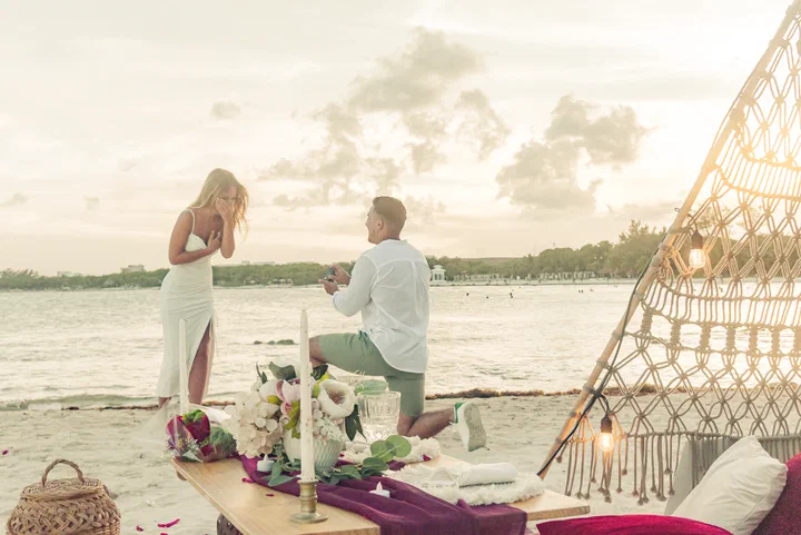 Hombre arrodillado pidiendo matrimonio junto a un picnic de macramé en una playa de Tulum