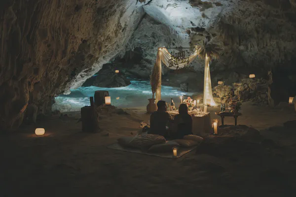 Montaje de cena romántica dentro de una cueva de cenote con una pareja sentada en una mesa bellamente decorada, letrero de 'Feliz Cumpleaños' arriba