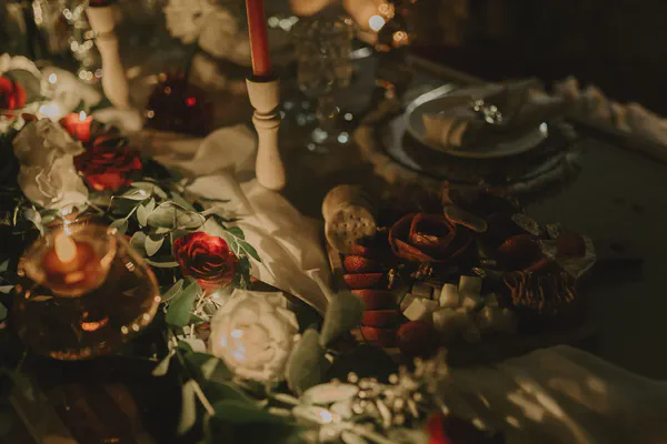 Primer plano de una tabla de charcutería con fresas, quesos y galletas junto a rosas rojas y velas