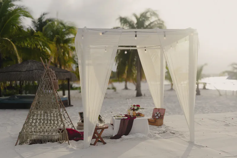 Montaje de cena romántica en la playa bajo un dosel blanco con cortinas suaves, canasta de picnic y arreglos florales