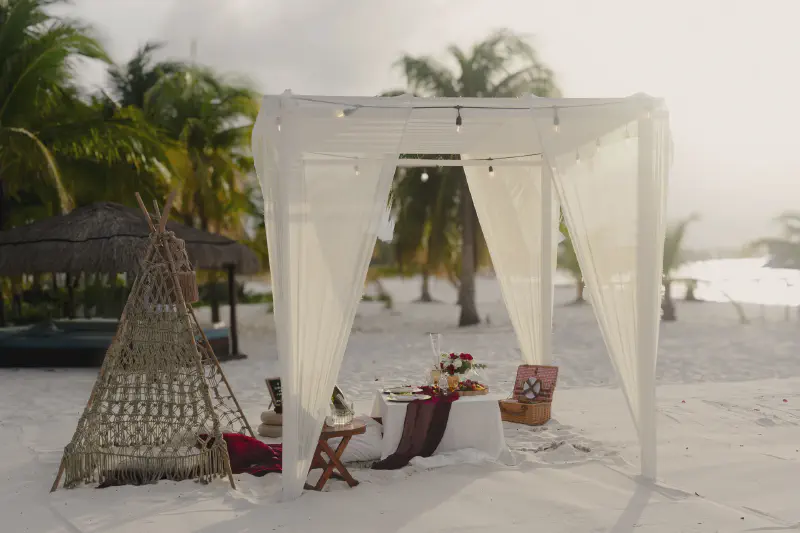 Romantic beach dinner setup under a white canopy with soft flowing curtains, picnic basket, and floral arrangements