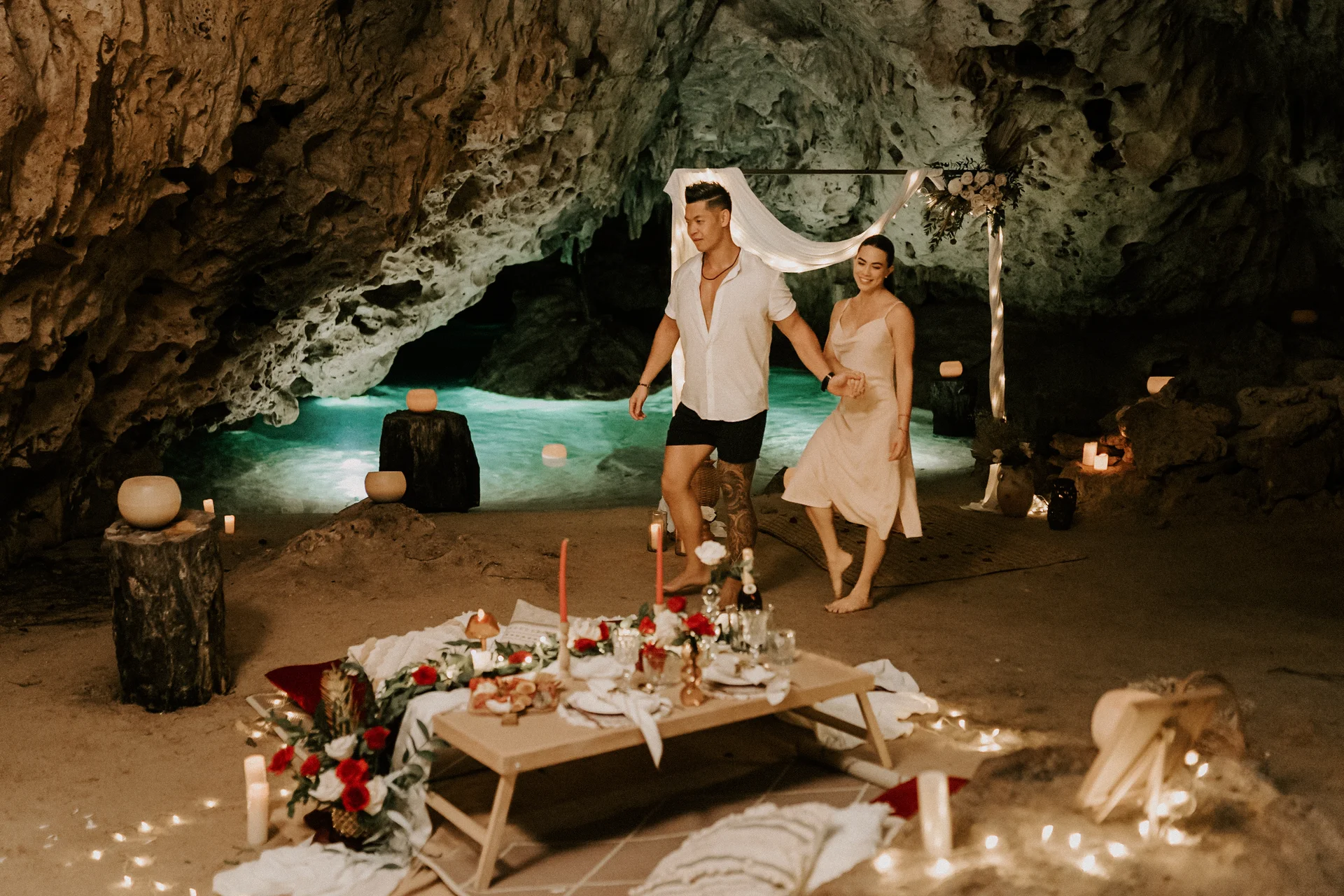 Couple walking hand-in-hand toward a romantic dinner setup inside a cenote cave, lit with candles and adorned with flowers