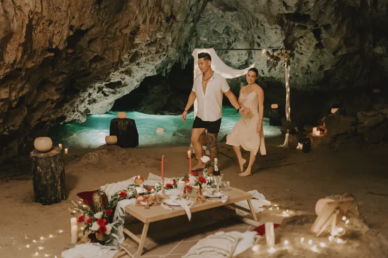 Couple walking hand-in-hand toward a romantic dinner setup inside a cenote cave, lit with candles and adorned with flowers