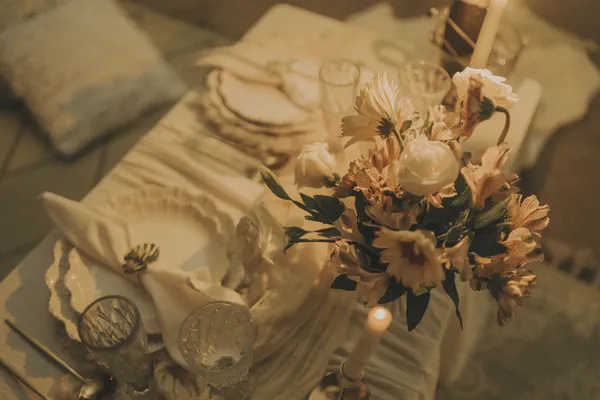 Primer plano de una mesa elegantemente decorada con copas de cristal, platos finos y un ramo de flores rosadas y blancas, iluminada por velas