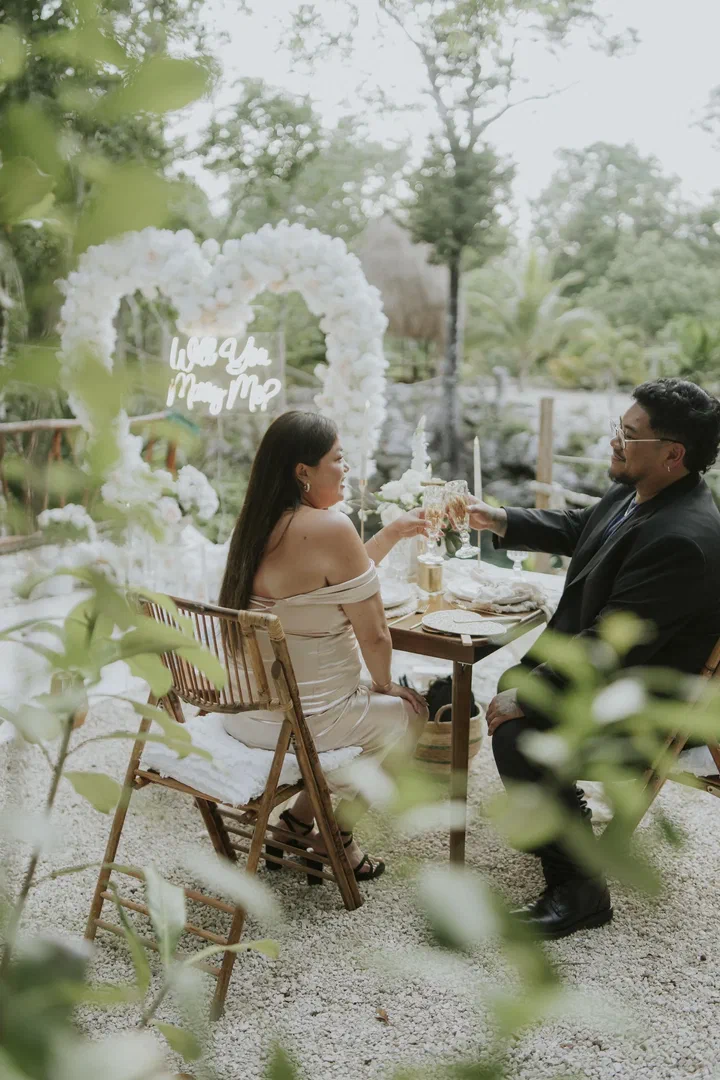 A couple raises a toast at a table for two in the jungle, with a white floral heart behind them.