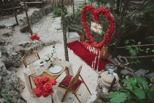 Outdoor marriage proposal setup: a red rose heart arch with 'Will You Marry Me?' neon sign. A red velvet runner, lit by candles and lanterns, leads to the arch. A dinner table for two, with white flowers and champagne, is set nearby in a natural, rocky landscape with lush greenery, ideal for an engagement.