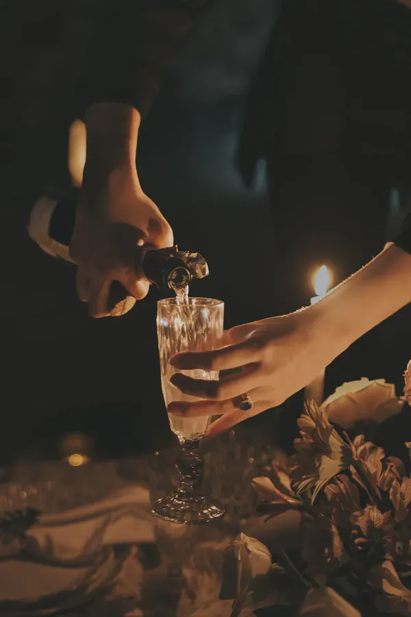 Close-up of hands pouring sparkling champagne from a bottle into a faceted glass flute, illuminated by the warm, golden glow of candlelight. The intimate, dark setting includes a hint of a floral arrangement on a table, with one hand wearing a ring, evoking a romantic or celebratory mood.