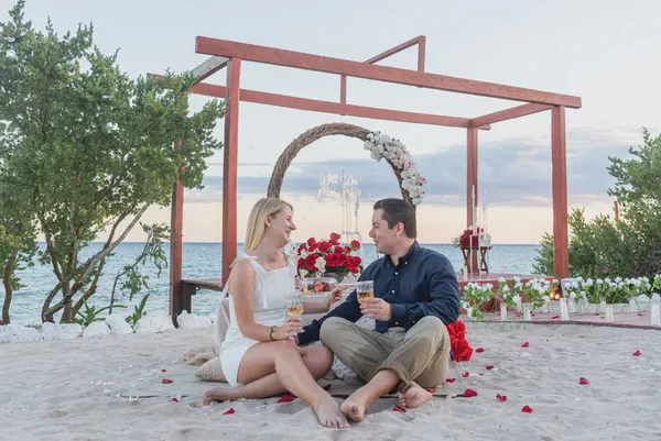 A happy couple sits barefoot on a sandy beach enjoying a romantic proposal dinner. Behind them, a flower-decorated wooden arbor frames a 'Will You Marry Me?' neon sign. Champagne, candles, and red rose petals create an intimate engagement setting by the ocean at twilight, capturing a joyous moment.