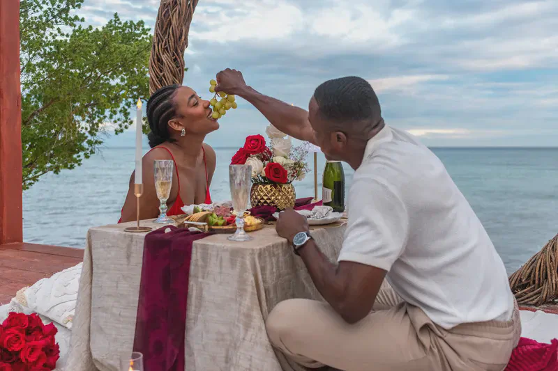 Una pareja disfruta una cena romántica al aire libre junto al océano al atardecer. El hombre alimenta con uvas a la sonriente mujer con vestido rojo. Su mesa tiene copas de champán, tabla de charcutería y ramo de rosas rojas, con el mar azul y cielo nublado al fondo.