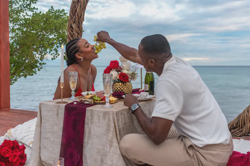 A couple enjoys a romantic outdoor dinner by the ocean at sunset. The man feeds grapes to the smiling woman in a red dress. Their table features champagne flutes, a charcuterie board, and a bouquet of red roses, set against the calm blue sea and cloudy sky.