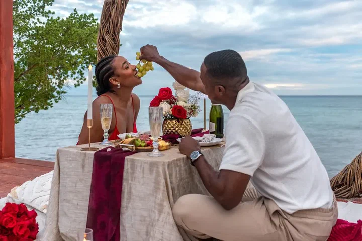 A couple shares grapes at a table set with champagne and red roses by the sea.