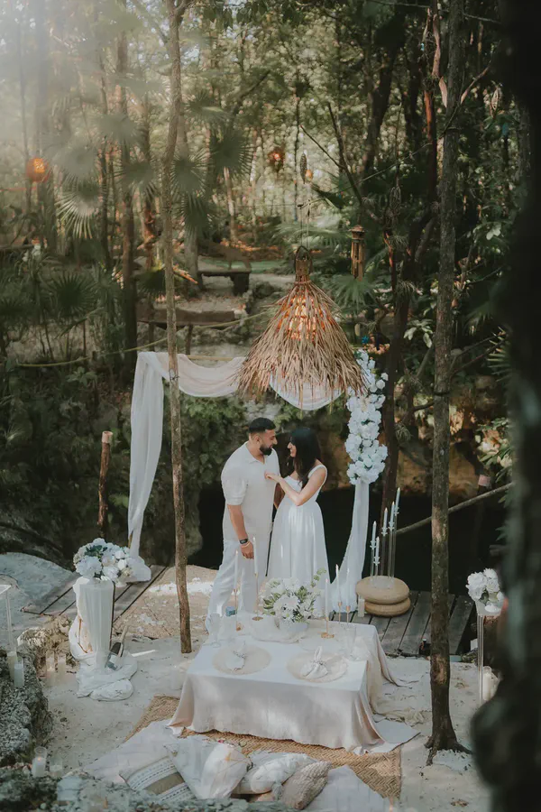 A romantic intimate dinner setup in a lush tropical jungle for a couple in white outfits. They gaze lovingly at each other by a low table adorned with white flowers, candles, and champagne. White drapes, a large woven lamp, and floor pillows complete the elegant, dreamy ambiance, perfect for a proposal or elopement.