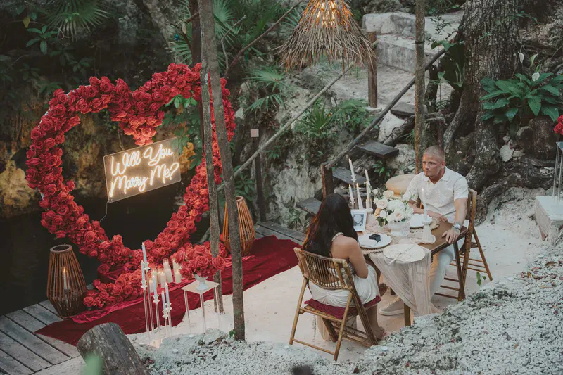 A romantic outdoor marriage proposal setup with a couple seated at a candlelit table in a lush, natural setting, possibly a cenote or jungle. Behind them, a large heart-shaped arch adorned with vibrant red roses features a glowing &lsquo;Will You Marry Me?&rsquo; neon sign. The intimate scene includes a red velvet carpet and white gravel, creating a magical atmosphere.