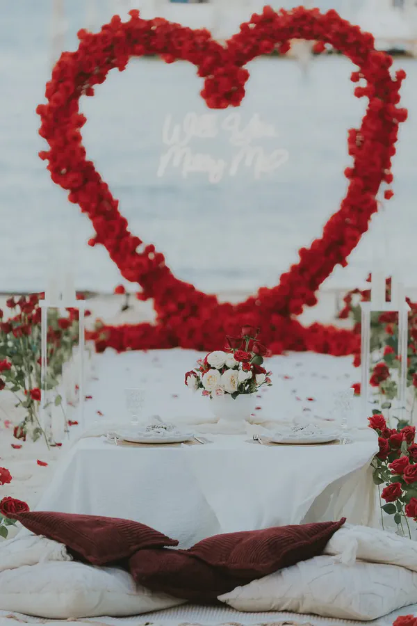 A romantic outdoor marriage proposal setup by the water. A large red rose heart arch frames a glowing 'Will You Marry Me?' neon sign. An intimate low table for two is set with white linen, elegant plates, and a beautiful centerpiece of red and white roses. Cushions on the ground, covered in scattered red rose petals, complete this enchanting engagement scene.