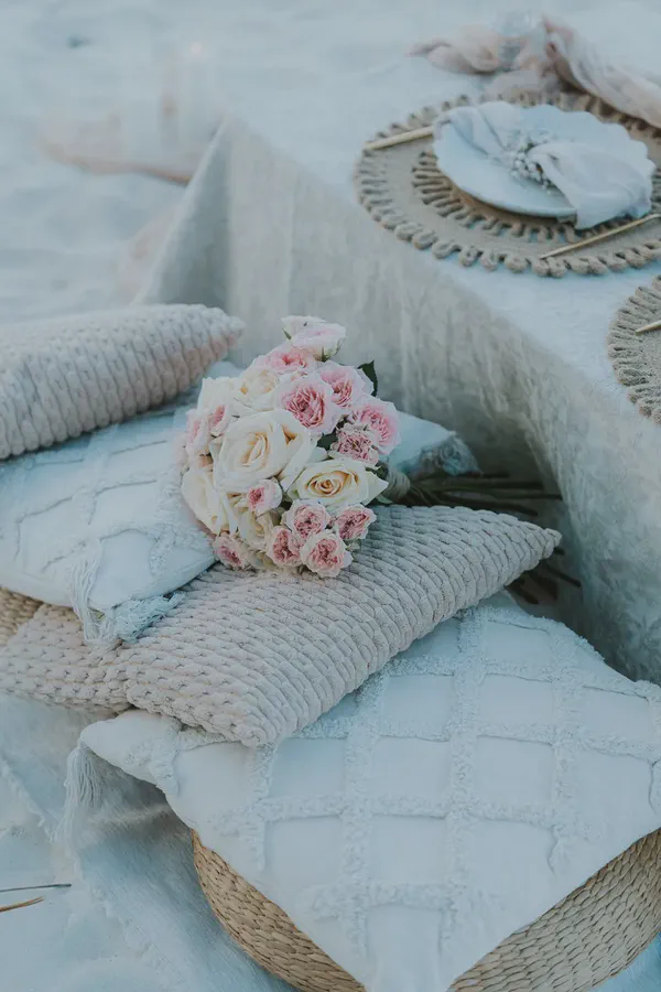 A romantic outdoor picnic setup features a bouquet of soft pink and creamy white roses on textured beige and white decorative pillows. In the background, a table is set with a woven placemat, white plate, and light napkin on a pastel-colored tablecloth, all on light sand. The scene evokes a tranquil, elegant, and intimate atmosphere.