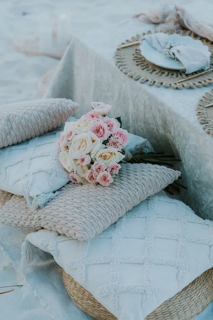 A bouquet of pink and cream roses on textured cushions beside a table set on the sand.