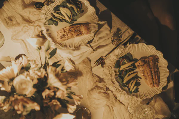 Overhead view of a romantic dinner table for two. Each plate features a perfectly cooked salmon fillet with a side of fresh green salad, topped with sliced fruit and red onion. Golden cutlery rests beside white scalloped plates on a light-colored, textured tablecloth, illuminated by warm, golden light, with blurry flowers in the foreground.