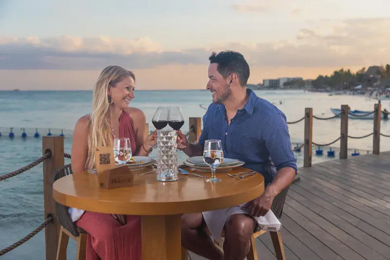 A romantic couple enjoying an intimate dinner on a wooden boardwalk overlooking the ocean at sunset. The man in a navy shirt is serving the woman in a red dress, who is seated at a beautifully set table with champagne and elegant tableware. The warm evening light creates a magical atmosphere with the ocean and lounge chairs visible in the background.