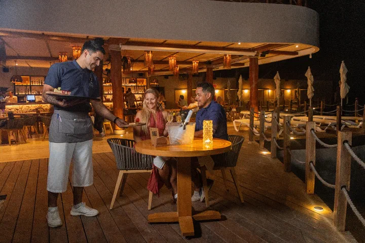 A waiter serves a couple at a candlelit table on an ocean-view restaurant deck at night.