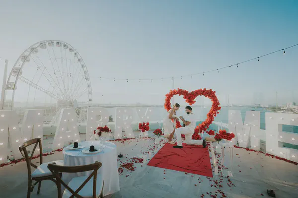 Romantic rooftop marriage proposal. A man kneels to a woman before illuminated 'MARRY ME' letters and a red rose heart arch. Red petals and a runner are on the floor. A Ferris wheel and city view are in the background under a blue sky.