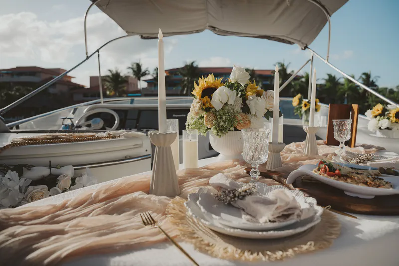 Luxurious dining table set on a yacht. Features a vibrant floral centerpiece of sunflowers and white roses, tall white candles, crystal glasses, and elegant plates. A peach runner covers the table. The sunny coastal background with palm trees suggests a romantic, tropical event.