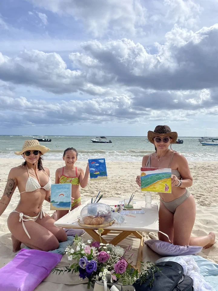 Two women and a girl on the beach hold up their paintings of the sea beside a low table with snacks and flowers.