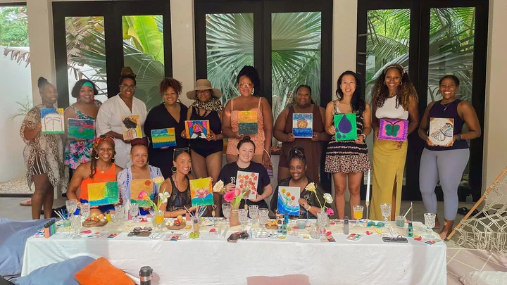A large group of women stand behind a long table and hold up the canvases they painted.