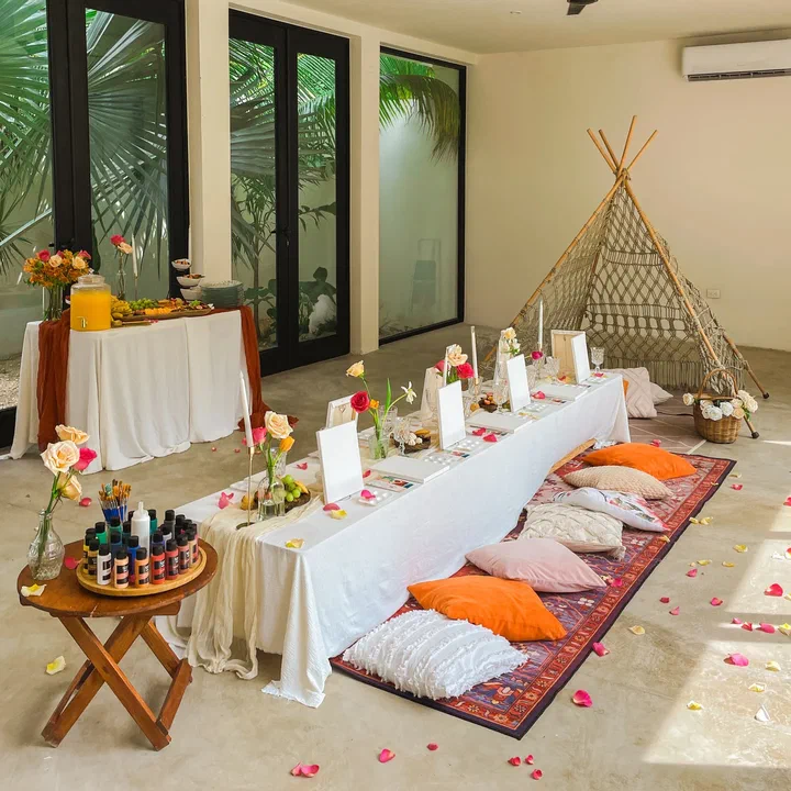 A long low table set with blank canvases, flowers and paint, with floor cushions and a macramé teepee, in a bright room with tall windows