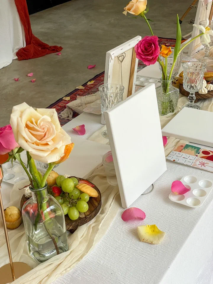 A blank canvas on a small easel beside roses, grapes and a paint palette on a white tablecloth.