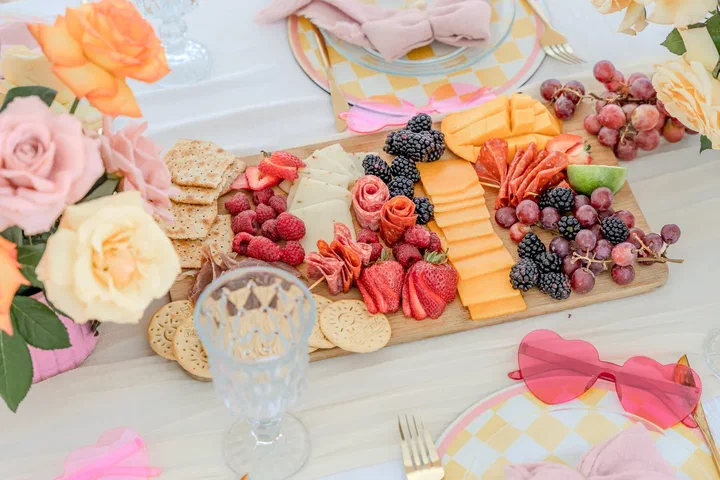 Charcuterie board with berries, cheese and grapes beside pink and peach roses, crystal glasses and heart-shaped sunglasses