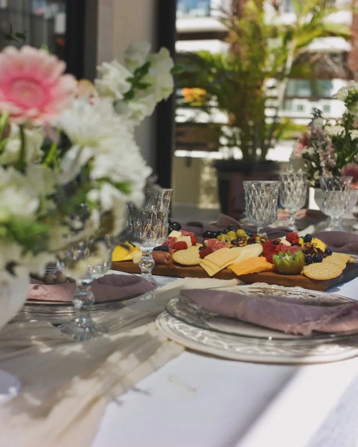 Una mesa con copas de cristal, flores rosas y blancas y una tabla de charcutería con quesos y fruta, junto a una ventana.