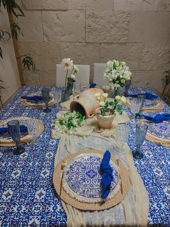 Una mesa con mantel estampado en azul y blanco, platos a juego sobre bajoplatos tejidos, servilletas azules y flores blancas en macetas de barro.