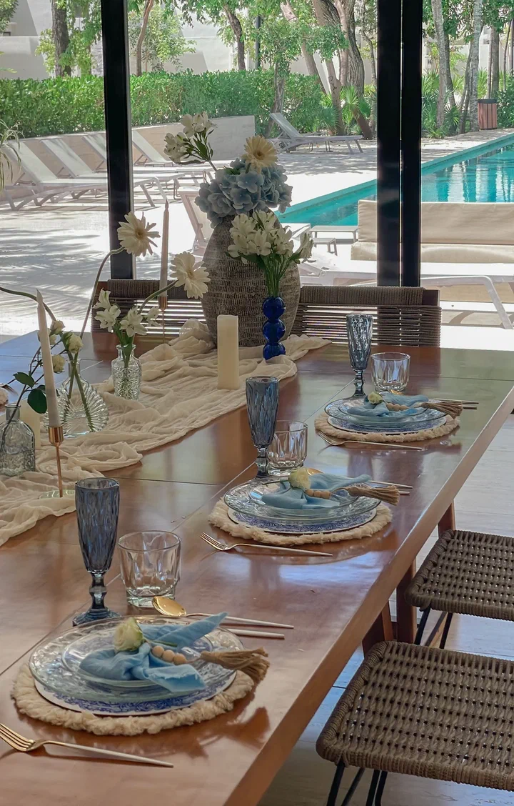 Una mesa de madera junto a una alberca con copas azules, servilletas azul claro sobre bajoplatos tejidos e hortensias azules.