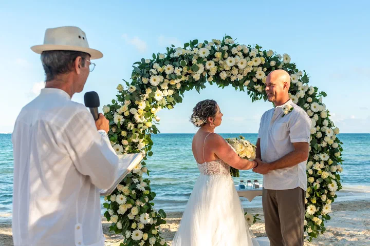 Una pareja se toma de las manos bajo un arco de flores blancas junto al mar mientras habla el oficiante.