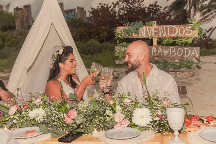 Una pareja brinda en una mesa con flores en la playa junto a un tipi blanco y un letrero de bienvenida.