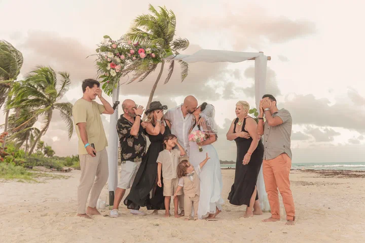 Familia y amigos ríen alrededor de una pareja bajo un arco adornado con flores en la playa.