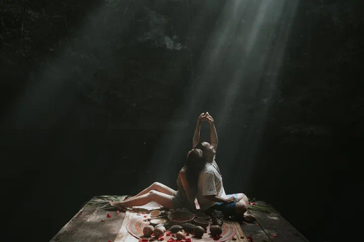 Una pareja sentada en una plataforma de madera en un cenote oscuro; uno de ellos alza la mano hacia un rayo de sol.