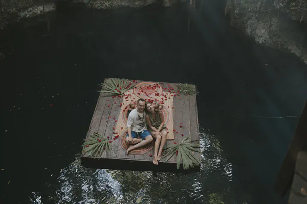 Aerial view of intimate elopement ceremony platform floating over crystal clear cenote waters in Tulum, decorated with palm fronds and rose petals for spiritual wedding