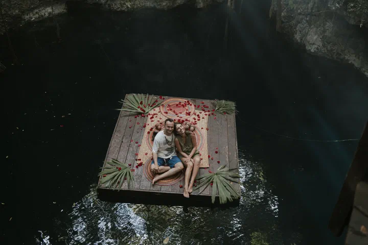 Vista desde arriba, una pareja recostada en una plataforma de madera con pétalos que flota sobre el agua oscura de un cenote.