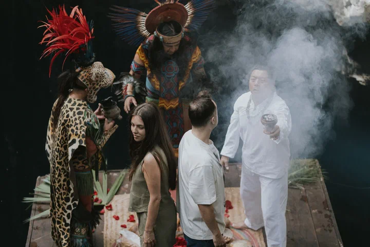 Un hombre vestido de blanco bendice a una pareja con humo de copal mientras danzantes mayas con penachos de plumas los acompañan en un cenote.