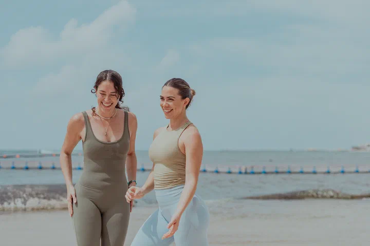 Two friends in activewear laugh together as they walk on the beach.