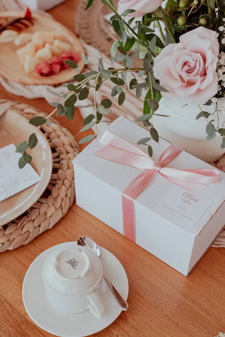 A white gift box tied with pink ribbon beside a coffee cup and pink roses on a wooden table.