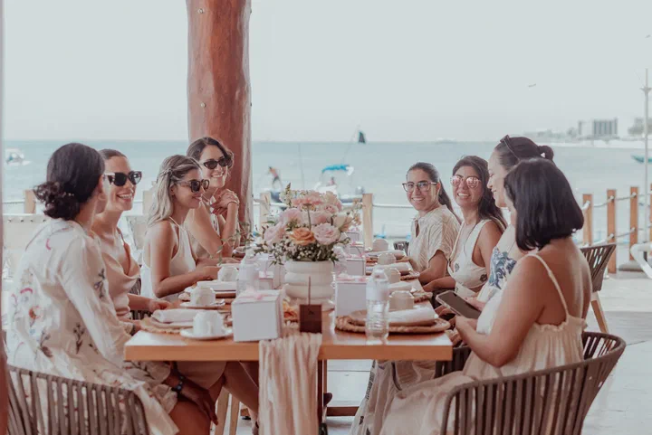 A group of friends laughs around a long brunch table with roses under a palapa by the sea.
